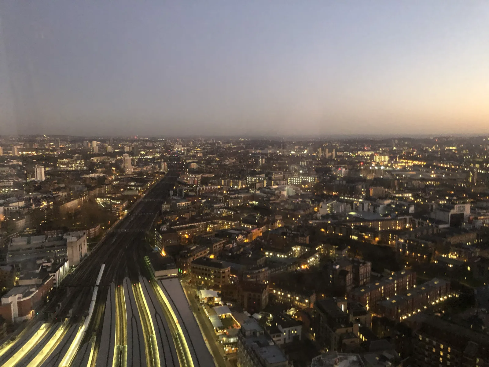 A high aerial view of a city at dusk, railway tracks fanning out from a station below and splitting into a dozen glowing lines as the lights spread to the horizon.